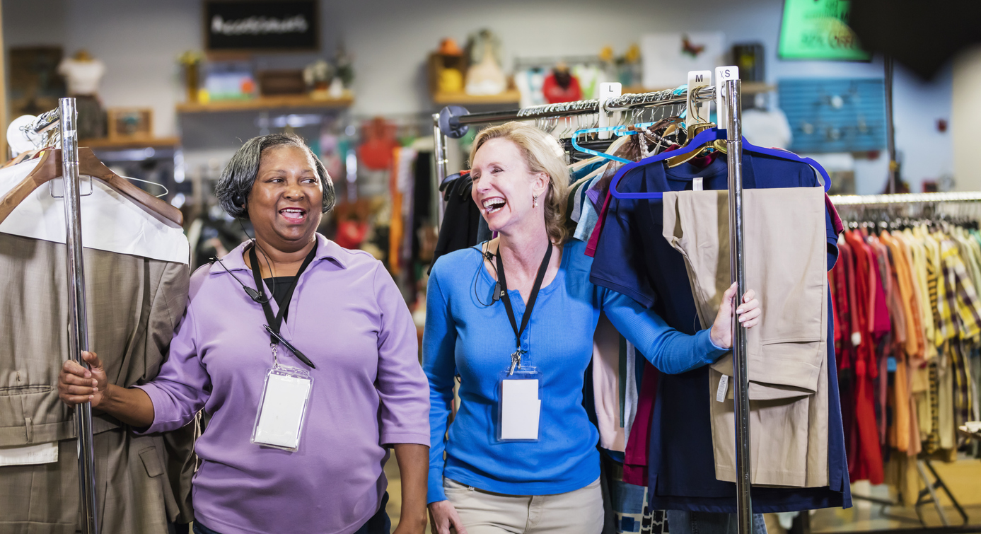 Two Thrift Store Volunteers Wheeling Racks of Clothing to the Sales Floor from the Backroom